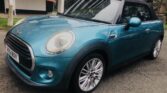 A teal-colored MINI Cooper convertible is parked on a residential street. The car, featuring a black soft-top roof and silver alloy wheels, proudly displays a European license plate at the front. In the background, trees, a building, and patches of grass can be seen under an overcast sky.
