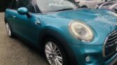 A teal MINI Cooper convertible is parked on a residential street. The front of the car faces the camera, showcasing its distinctive round headlights and chrome grille. The car has a black fabric roof in the down position. Several other parked vehicles and residential buildings are visible in the background.