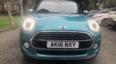 Front view of a teal MINI Cooper convertible parked on a street. The car has its headlights on and a dark interior visible through the windshield. Trees and other vehicles can be seen in the background. The license plate reads "AK16 NXY.