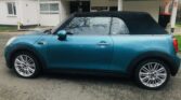 A teal MINI Cooper convertible with a black soft top is parked on a residential street in front of a beige brick and white-paneled apartment building. The car, facing left and visible from the side, features silver alloy wheels. Behind it, a sign reads "Woodhall House.