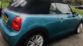 A teal MINI Cooper convertible with a black soft top is parked on the side of a road. The car has a UK license plate reading "AX19 VKX." Bare trees and a grassy area can be seen in the background. The pavement is slightly wet, suggesting recent rain.