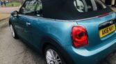 A blue MINI Cooper convertible with a black soft top is parked on a paved street near a park with trees and children in the background. The car, boasting prominent rear lights and alloy wheels, has a UK registration plate reading AK16 NXY.