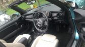The image shows the interior of a MINI convertible car viewed from the passenger side. The car features off-white leather seats, a black dashboard, and a black steering wheel with a white logo at the center. The gear lever, air vents, and entertainment display are visible. Green outdoor scenery can be seen through the open top.