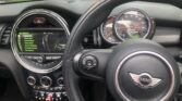 The interior of a MINI Cooper car features the signature steering wheel with the MINI logo, a central control panel with a digital display, various control buttons, and two air vents. The dashboard proudly showcases speedometer and tachometer gauges. A street and greenery are visible through the windshield.