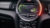 Interior view of a MINI car showing the dashboard with a central circular display screen. The screen currently displays a driving mode labeled "SPORT Mode" with graphics and a green line. Below the screen are various control buttons and dials, and the car's gear stick is visible at the bottom.