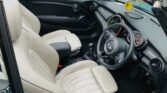 Interior view of a MINI convertible car with beige leather seats. The steering wheel is on the right side, indicating a right-hand drive vehicle. The dashboard features several gauges and a circular infotainment system. A yellow air freshener hangs from the rear-view mirror, while "LD07 RXM" is visible in the background.