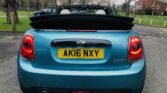 A light blue MINI Cooper convertible is parked on a residential street. The car's roof is down, and the rear license plate reads "AK16 NXY." This MINI features round taillights and a compact design. In the background, there are bare trees, green grass, and houses on a cloudy day.