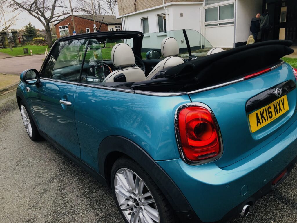 A parked blue MINI Cooper convertible with a black soft top in a lowered position stands on a residential street. The car's registration plate reads "AK16 NXY." Houses, greenery, and a driveway provide the backdrop, while the convertible’s tan and black interior is visible.