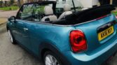 A parked blue MINI Cooper convertible with a black soft top in a lowered position stands on a residential street. The car's registration plate reads "AK16 NXY." Houses, greenery, and a driveway provide the backdrop, while the convertible’s tan and black interior is visible.