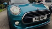 A blue MINI Cooper with its headlights on is parked on a residential street. The car's license plate reads "AK16 NXY." In the background, other cars are parked near a multi-story brick building surrounded by tall trees. The photo is taken from a low angle, emphasizing the front grille and headlights.