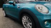 A teal MINI Cooper convertible is parked on a road, with its top down. The car features modern halogen headlights and sporty alloy wheels. Trees and a residential building are visible in the background, slightly out of focus. The image is taken from a low, diagonal front angle.