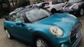 A teal MINI convertible is parked in a lot with the top down. Adjacent to it are a white SUV, a gray MINI, and several other vehicles. Residential buildings and trees are visible in the background. The car's lights are on, and it features chrome detailing.