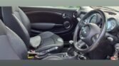 The image showcases the interior of a compact MINI, with visible features including dark seats, a steering wheel adorned with a keychain, and a gear shift in the central console. The dashboard houses round air vents and a speedometer, with a mixture of dark and metallic accents throughout.