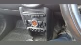 The image showcases the interior of a MINI, emphasizing the central console area. It features a round display and several control buttons, including what appears to be air conditioning or radio controls. The floor mat beneath is visible, along with part of the steering wheel and the driver’s foot wearing a black shoe.