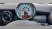 The dashboard of a MINI car shows a close-up of its large circular speedometer with a digital screen below it. The display reads "Range 5 miles," "MPG 19.5," and "045011 miles." A small section of the air vent and the carbon fiber trim on the dashboard are visible to the left.