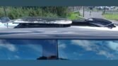 Close-up of a MINI roof with a damaged and partially detached black sunroof panel. The sky is reflected in the vehicle's rear windows, and the background includes greenery and metallic fencing, indicating an outdoor setting. The image's top and bottom edges fade into different shades of blue.