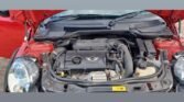 The image shows the engine compartment of a red MINI Cooper with its hood open. The engine and surrounding components are visible, including the MINI logo on the engine cover. Various hoses, wires, and parts of the car's front structure can also be seen. The background is blurred out.