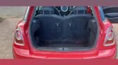 The image shows the rear of a red MINI with its hatchback trunk open. The trunk space is empty, revealing the back of the rear seats and part of the car's stylish interior. The background features a gradient from teal at the top to red at the bottom.