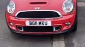 A red MINI Cooper with a license plate reading "BG11 MRU" is parked half on the pavement and half on the road. The car is facing the camera. A street with houses, trees, and parked cars is visible in the background. The sky is slightly overcast.