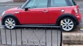 A red MINI Cooper is parked on a residential street in front of a house with a red-tiled roof. The car sits next to a low brick wall with a decorative metal gate, blending into the charming neighborhood. The background includes trees, bushes, and other houses under an overcast sky.