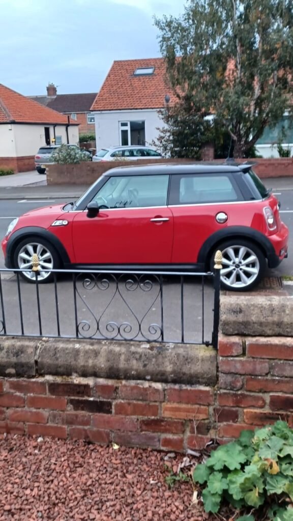 A red MINI Cooper is parked on a residential street in front of a house with a red-tiled roof. The car sits next to a low brick wall with a decorative metal gate, blending into the charming neighborhood. The background includes trees, bushes, and other houses under an overcast sky.