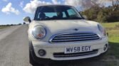 A white MINI Cooper with the license plate "MV58 EGY" is parked on the side of a rural road. The background features a partly cloudy blue sky and green shrubs. The car faces the camera, showcasing its front grille and headlights. The road runs off into the distance on the left.