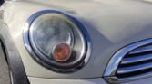 Close-up of the front left headlight of a white MINI Cooper. The headlight features a clear lens with chrome trim and visible bulbs inside, including an orange indicator light. Part of the front grille, also accented with chrome, is visible on the right side. The car is parked outdoors.