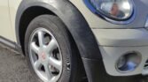 Close-up of the front left side of a white MINI Cooper car. The image clearly shows a silver alloy wheel with a black tire, part of the headlight, and the front bumper with a small circular fog light. The car's body is clean and well-maintained, and the pavement is visible beneath the car.