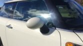 Close-up of a white MINI's side, showing the driver's door, side mirror, and a portion of the front windshield. The car is outdoors, and reflections of the sky with clouds and surrounding greenery are visible on the windows. The side mirror is white, matching the car's color, and there is a small amber indicator light on the side.