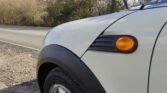 Close-up of the front left corner of a light-colored MINI parked on the side of a road. The car features a black fender and side marker light. The background shows a tree-lined road and a clear sky with some clouds. The tire and part of the wheel are visible.