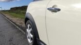 Partial rear side view of a parked white MINI Cooper on a countryside road. The car's rear right tire and side door handle are clearly visible. The background shows a hedge, grassy verge, and a partly cloudy sky. The road is paved and runs parallel to the hedge.