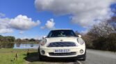 A white MINI Cooper is parked on the roadside near a calm lake under a bright blue sky filled with puffy white clouds. The car's front license plate reads "MV58 EGY." The scene includes green grass, bare trees, and a rural, peaceful backdrop.