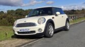 A white MINI Cooper with the license plate "WV58 EGY" is parked partially on a grassy area and partially on an asphalt road, next to a wooden fence. The car is bathed in sunlight, with trees, a lake, and a partly cloudy sky in the background.