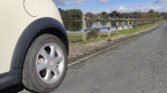 A white MINI is parked on the roadside, with a focus on its rear wheel. Beside it, there's a narrow, paved pedestrian path and a wooden fence separating the path from a small lake that reflects the blue sky and scattered clouds. In the background are trees and grassy areas.