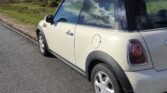 A cream-colored MINI Cooper is parked on the side of a paved road. The road is bordered by green grass and bushes. The MINI reflects clouds in the sky on its windows and roof. The car's fuel cap and rear brake lights are visible from this angle.