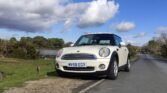 A white MINI Cooper with a license plate reading "MV58 EGY" is parked along a rural road. The road is bordered by grass and trees, and a small body of water is visible in the background. The sky above is partly cloudy with patches of blue.