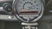 Close-up of a MINI car dashboard featuring a central speedometer reaching up to 160 mph. Below it, the digital display shows the time as 11:49 and options for Vol and FUK. Beneath the digital readout are buttons labeled with symbols and a row of toggle switches.