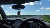 View from inside a MINI showing the dashboard and steering wheel. An air freshener hangs from the rear-view mirror. The shot, oriented through the windshield, captures a road stretching ahead with green bushes on either side and blue sky with scattered clouds above.