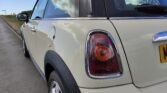 Close-up of the rear side of a light-colored MINI Cooper with a black roof and distinctive taillights. The background shows a road stretching alongside greenery and a blue sky with scattered clouds. The car's right rear section is prominently visible.