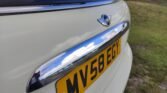 Close-up of the rear of a beige MINI Cooper car, featuring a chrome-finished handle and the MINI logo above it. The license plate reads "MV58 EGY" and is yellow with black text. The background shows grass and a blurry tree. The sky is reflected on the car's chrome handle.