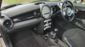 Interior of a MINI showing the front seats, dashboard, and steering wheel. The dashboard features a large, central speedometer, with air vents on either side. There are various controls below the speedometer, including the climate controls and radio. The gear stick and cup holders are also visible.