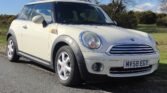 A cream-colored MINI Cooper with a UK license plate reading "MV58 EGY" is parked on the side of a rural road. The car has a silver grille, circular headlights, and black trim. In the background, there are green fields, bushes, and a leafless tree against a clear blue sky.