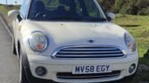 A white MINI Cooper is parked on the side of a paved road next to a grassy area. The front view of the car shows a silver grille, round headlights, and a license plate with the registration "MV58 EGY." Trees and bushes are visible in the background under a partly cloudy sky.