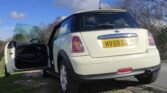 A white MINI Cooper with a UK license plate "MV58 EGB" is parked on the side of a road. The driver's side door is open, and the car rests in a grassy area surrounded by trees with a cloudy sky in the background. The scene depicts a calm, sunny day.