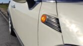 Close-up of the front left side of a white MINI on a road. The image highlights the side mirror, orange turn signal, and part of the windshield. The car's design includes a black accent with grooves next to the turn signal. Sunlight reflects off the car's surface, creating a glare.