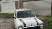 A white classic MINI Cooper with black racing stripes on the hood and a Union Jack painted on the roof is parked in front of a closed beige garage door. The car's license plate reads "J29 RJB." Cloudy sky and some grassy patches with gravel are visible in the foreground.