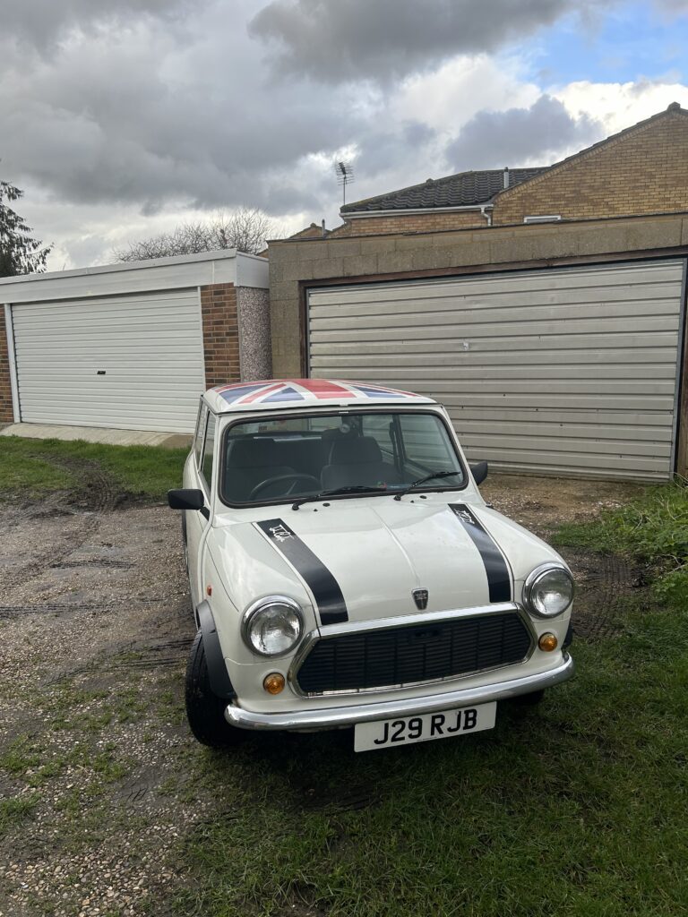 A white classic MINI Cooper with black racing stripes on the hood and a Union Jack painted on the roof is parked in front of a closed beige garage door. The car's license plate reads "J29 RJB." Cloudy sky and some grassy patches with gravel are visible in the foreground.