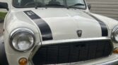 A classic white MINI Cooper is parked on a grassy area in front of a garage. The car features two black racing stripes on the hood, round headlights, and the MINI emblem on the grill. The license plate reads "J29 RJB." The background includes a wooden garage and parts of neighboring buildings.