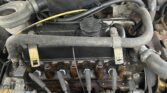 A view of a MINI car engine showing various components, including spark plug wires, an air filter, and connecting hoses. Visible wiring runs along the engine bay, with some signs of rust and wear on the metal parts. The overall appearance suggests an older, possibly vintage, MINI engine model.
