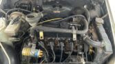 The image shows the engine bay of a MINI, with various components and connections visible. The focus is on the engine in the center, with numerous hoses, wires, and a yellow-labeled cylindrical part. The surrounding area includes the battery, coolant reservoir, and other mechanical parts, all slightly worn.